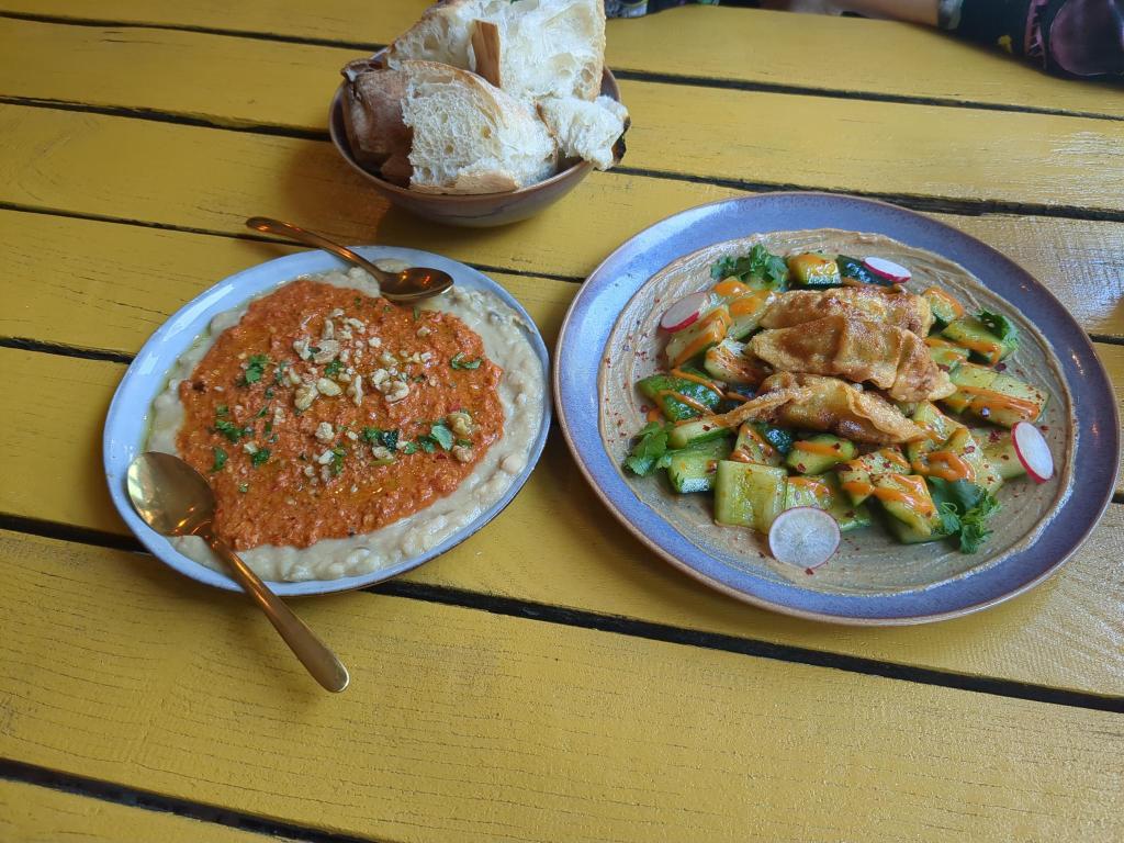 Picture of three plates of food on a wooden table from the Eden Restaurant in Saarbrucken. On the left, Muhammara with White Beans and on the right, Cucumber and Gyoza Salad.At the top of the image, a basket of white baguette
